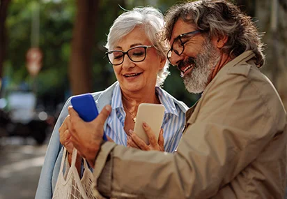 homem e mulher cerca de 60 anos mostrando celular um para outro em ambiente externo